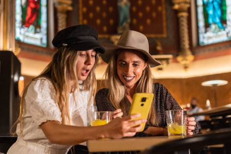 Two tourist friends looking on the phone at leisure activities in a modern cafeteria. Sipping an...