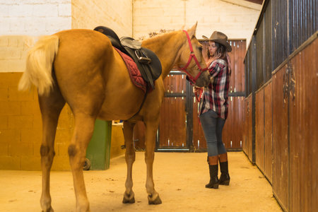 Caucasian cowgirl woman strolling with a horse in a stable, southern usa hats, pink plaid shirt and jeansの写真素材