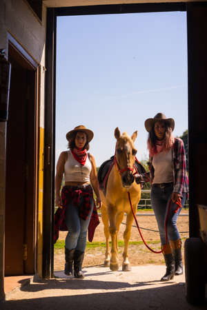 Two cowgirl women entering the stable with a horse on a horse, with South American outfitsの写真素材