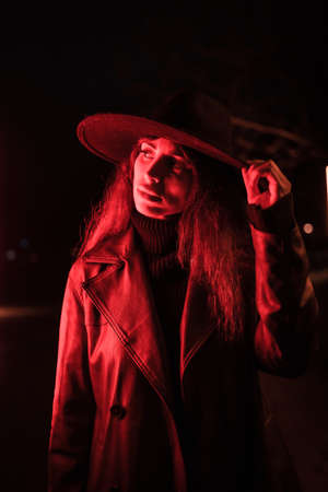 Girl in a hat in a portrait at night in a raincoat on a parcel. Illuminated with dim red light from the sideの写真素材