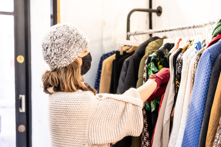 A customer with a protective mask looking at new collection clothes on a hanger, second-hand and...