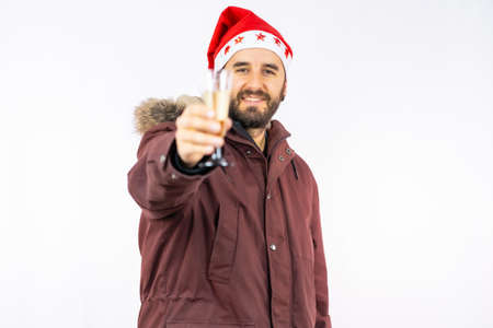 Young very happy Caucasian man with red Christmas hat toasting with a glass of champagne on a white background, dressed in a winter jacketの写真素材