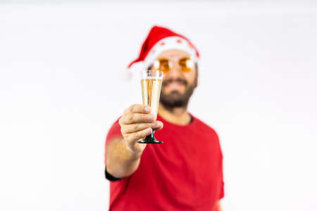 Very happy young Caucasian man with red Christmas hat toasting with a glass of champagne on a white background, dressed in a red t-shirt and sunglassesの写真素材