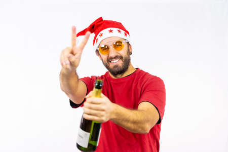 Young very happy Caucasian man with red Christmas hat toasting with a bottle of champagne on a white background, dressed in a red t-shirt and sunglassesの写真素材