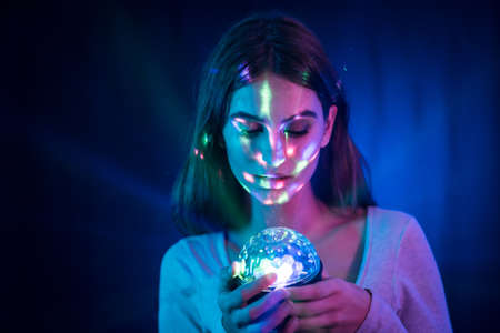 A young brunette Caucasian woman in a white t-shirt on a black background. Neon lights, illuminated with a disco light in handの写真素材