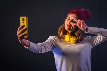 Young brunette caucasian with yellow headphones, white t-shirt and woolen hat on a black background, taking a selfie with mobile phoneの写真素材