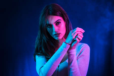 A young brunette Caucasian woman in a white t-shirt dancing in a nightclub with a black background. Neon lights, portrait looking at cameraの写真素材