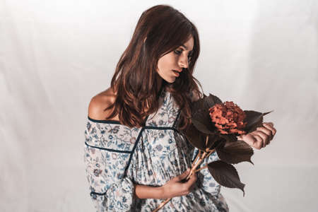 A young brunette Caucasian woman in a vintage floral dress on a white background. Portrait with a red flower in hand, with a sweet look in the studioの写真素材