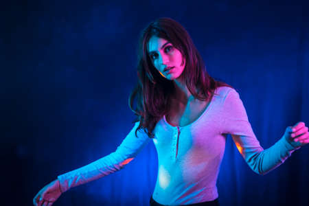 A young brunette Caucasian woman in a white t-shirt in a nightclub with a black background. Neon lights, dancing portraitの写真素材