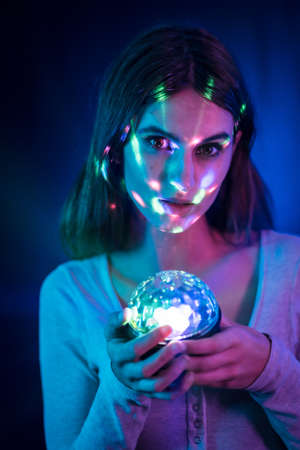 A young brunette Caucasian woman in a white t-shirt on a black background. Neon lights, illuminated with a disco light in hand, vertical photoの写真素材
