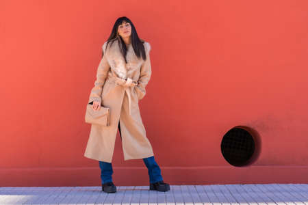 Posing of a smiling brunette model in a winter jacket leaning against a wall with a bag. Caucasian pretty a red backgroundの写真素材