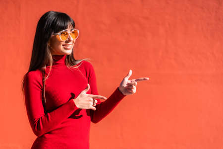 Brunette girl in a spring orange dress and sunglasses, pointing to an offer to the right. Copy and paste space. Caucasian pretty a red backgroundの写真素材