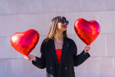 Caucasian brunette girl with a heart balloons on Valentine's day, sunglasses and red dress, On a gray background smiling and having funの写真素材