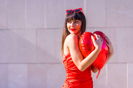 Brunette caucasian girl hugging heart balloon in valentine's day, sunglasses and red dressの写真素材