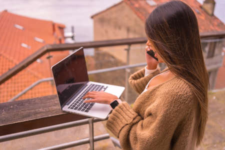 A young Latin woman with a white t-shirt and a brown wool sweater, teleworking on the terrace of a house by the sea, with her back to herの写真素材