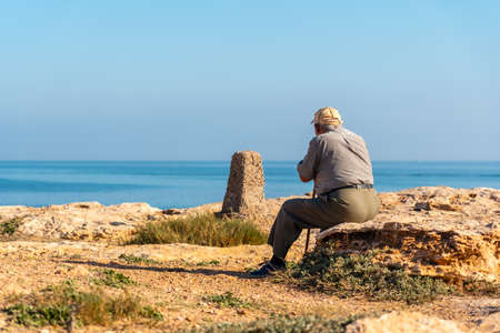 Pensive old man in beret sitting and with walking stick on the shore by the sea. Traditional lord of Spanish cultureの写真素材