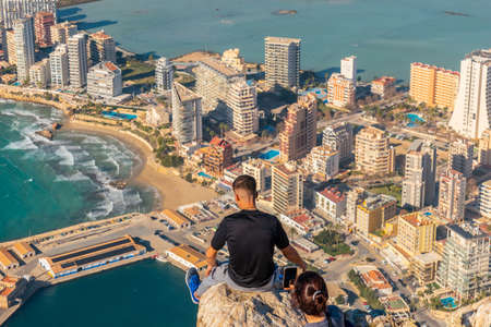 A young man sitting on top of the Penon de Ifach Natural Park with the city of Calpe in the background, Valencia. Spain. Mediterranean sea. View of the Cantal Roig and La Fossa beachの写真素材