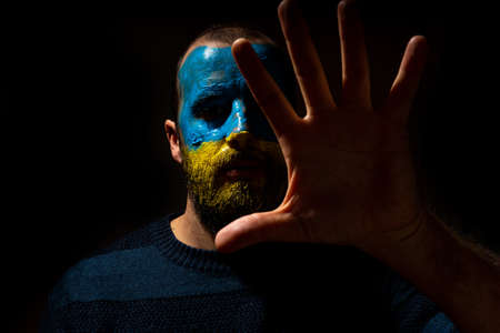 Stop war, conflict between Ukraine and Russia. Portrait of a young man with his face painted in the blue and yellow colors of the flag. Making the gesture to stop the warの写真素材