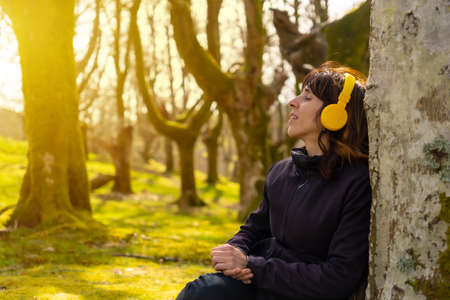 A girl listening to music with yellow headphones in the forest at sunset, sitting on a tree with her eyes closedの写真素材