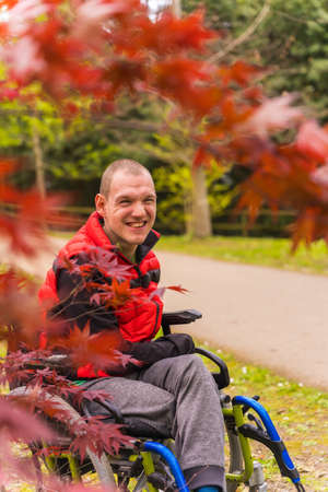 Portrait of a paralyzed young man in a red vest in a public park in the city. Sitting in the wheelchair next to red leavesの写真素材