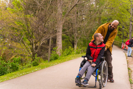 Paralyzed young man in the wheelchair being pushed by a friend in a public city park, strolling along a path having fun in springの写真素材