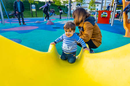 One year old Caucasian boy playing swings with his mother, playing in the park jumping on a yellow squeakerの写真素材
