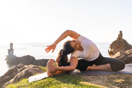 A Latin woman doing yoga exercises in nature by the sea, stretching next to a lighthouseの写真素材