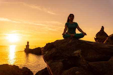 A woman doing meditation and yoga exercises at sunset next to a lighthouse in the sea, healthy and naturist life, outdoor pilates, ardha padmasanaの写真素材