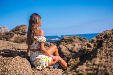 Portrait of a young woman in a white dress on the beach, sitting on some rocks looking at the seaの写真素材