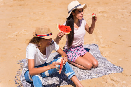 Two friends in summer on the beach eating a watermelon with the sea in the background, sitting on...