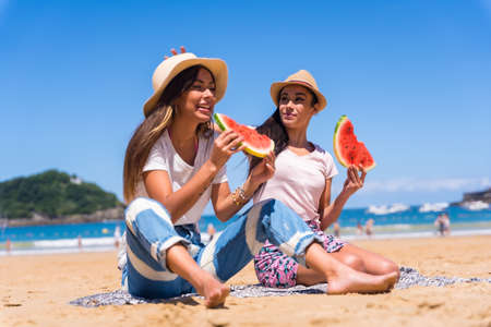 Two sisters in summer on the beach eating a watermelon, on vacation with the sea in the backgroundの写真素材