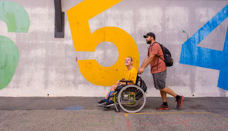 A disabled person in a wheelchair with a cement wall, walking with a friendの写真素材