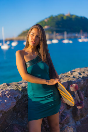 Portrait of a young Latin brunette woman with a skate wearing a green dress with the sea in the backgroundの写真素材