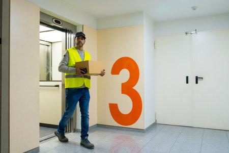 Young delivery man in protective uniform at online order delivery, going up elevator for home...