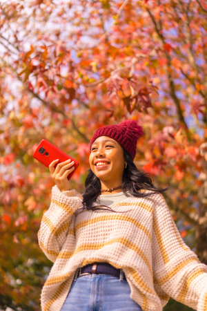 Asian girl in autumn with a mobile smiling sending a voice message in a forest of red leaves,...