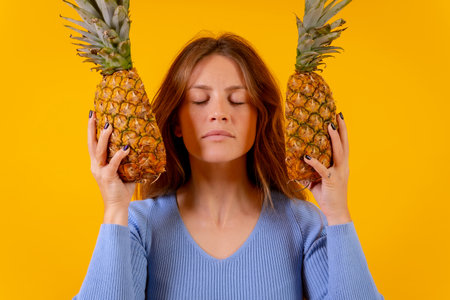 Woman with a pineapple in sunglasses, eyes closed with pineapple cut in half, yellow backgroundの写真素材