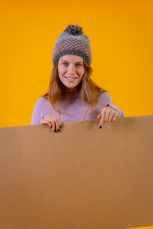 A woman in a wool cap holding and pointing at a cardboard sign on a yellow backgroundの写真素材