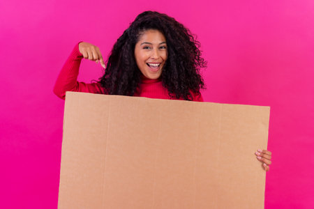 Curly-haired woman pointing at a sign on a pink background, studio shotの写真素材
