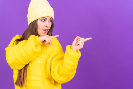 Attractive woman smiling pointing fingers at copy space on purple background, yellow wool sweaterの写真素材