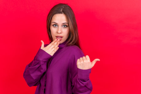 Attractive smiling woman pointing fingers at copy space on red background, surprised, good offerの写真素材