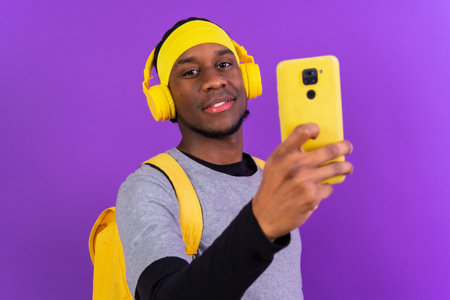 Black ethnic man with backpack and yellow headphones on a purple background, student concept, concept listening to music onlineの写真素材