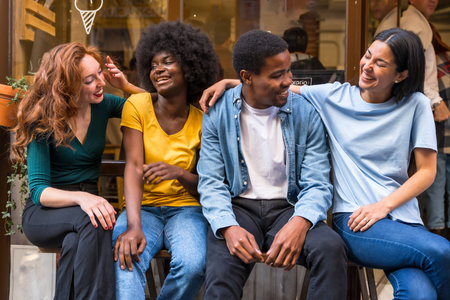 Portrait of multi-ethnic friends in a coffee shop sitting in the doorway of the shop window, embracing having funの写真素材