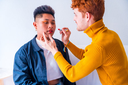 Beautiful gay couple putting on makeup painting their lips being romantic indoors at home, lgbt conceptの写真素材