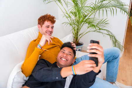 Beautiful gay couple being romantic indoors on the sofa, gay couple, taking a selfieの写真素材