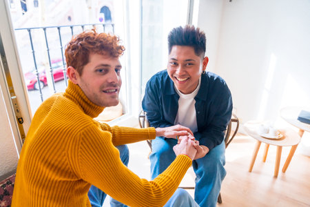Portrait of beautiful gay couple being romantic at home by the balcony holding hands, lgbt concept