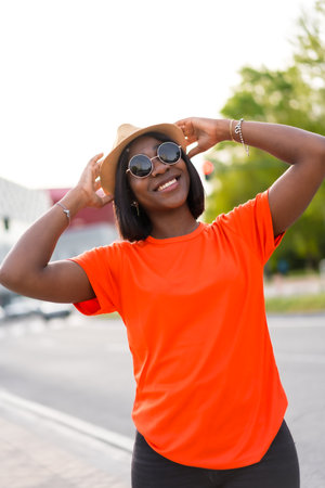 Smiling young black woman with sunglasses and hat, wearing orange t-shirt, enjoying summer in the city, lifestyle photosの写真素材
