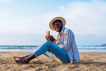 Portrait of a black ethnic man enjoy summer vacation at the beach eating a watermelon, Sitting on the sand smilingの写真素材
