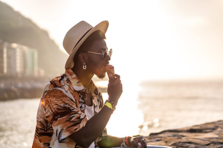 Sunset of a black ethnic man enjoy summer vacation on the beach eating an ice cream with a hatの写真素材