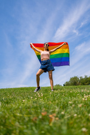 Caucasian brunette woman with a rainbow lgbt flag on the grass and blue sky backgroundの写真素材
