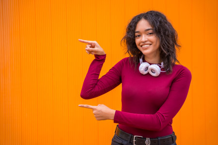 Portrait of latin woman with headphones smiling, technology concept on an orange background, pointing left, copy spaceの写真素材